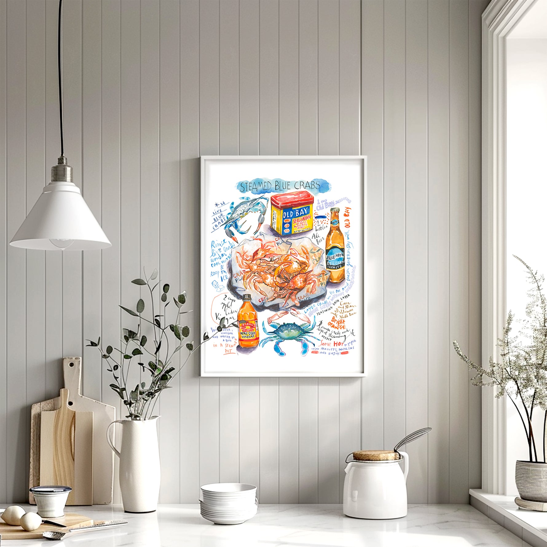 steamed blue crab recipe fine art print in watercolor featuring the ingredients and the finished dish, along with handwritten instructions in English. The framed poster hanging in a kitchen.