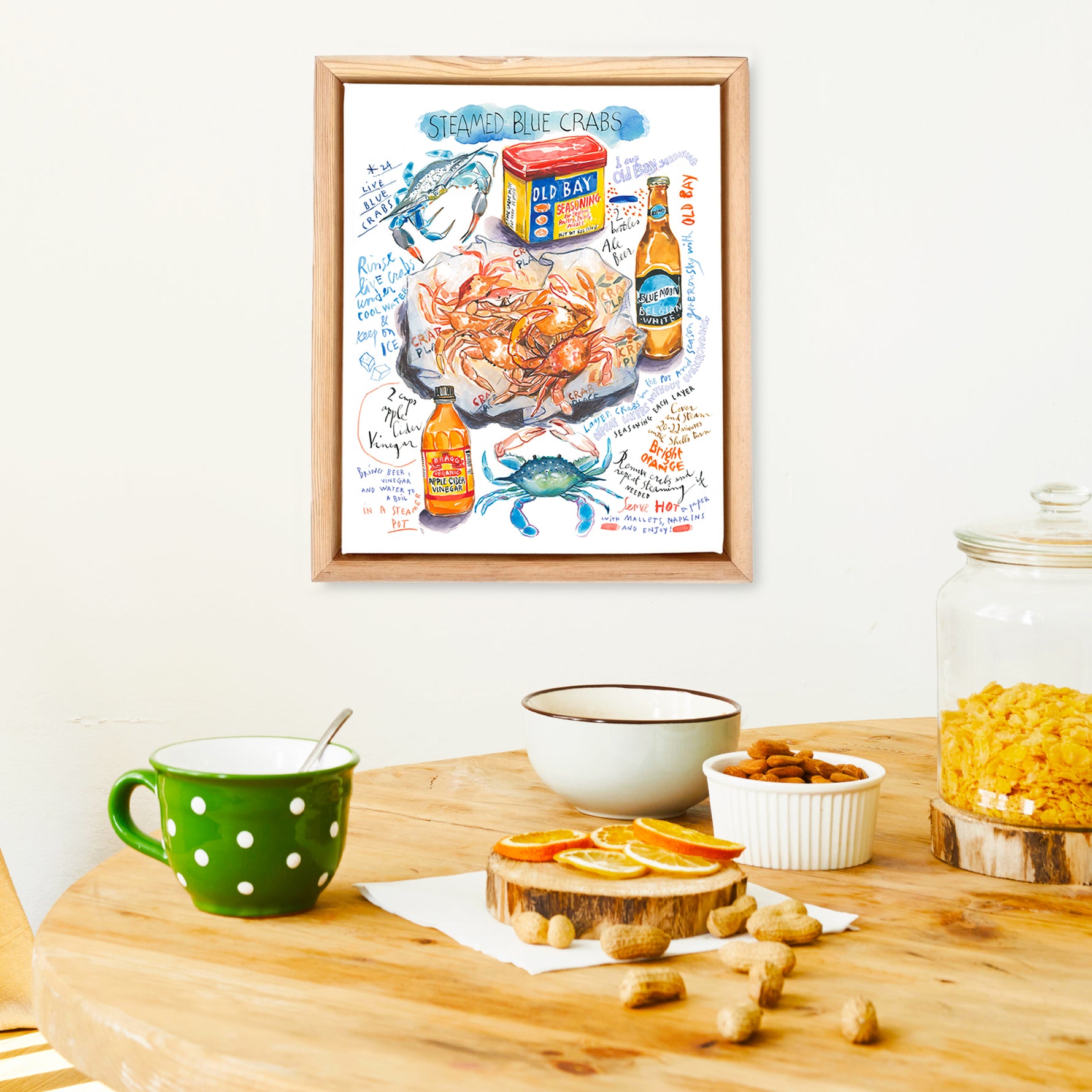steamed blue crab recipe fine art print in watercolor featuring the ingredients and the finished dish, along with handwritten instructions in English. The framed poster hanging in a dining space.
