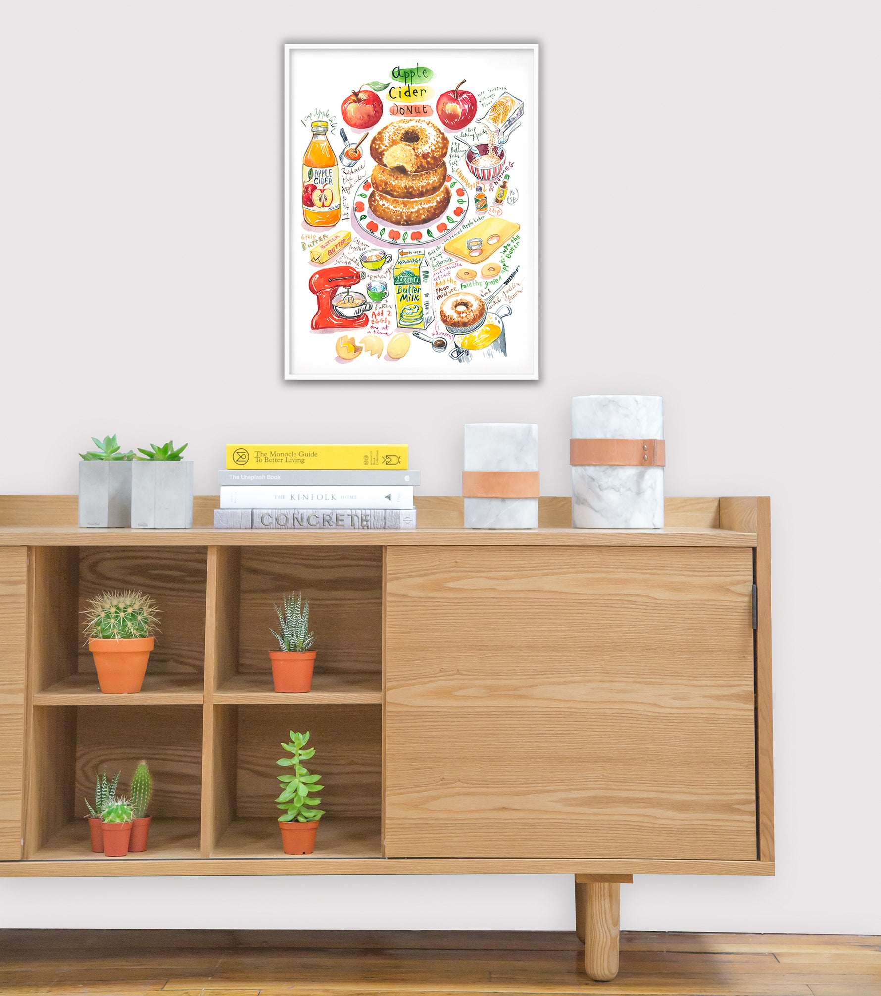 an apple cider donut recipe fine art print in watercolor featuring the ingredients and the finished dessert, along with handwritten instructions in English. The framed poster hanging in a home space.