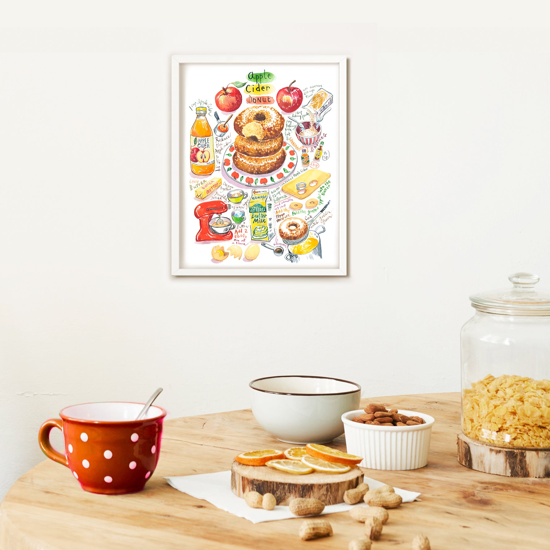 an apple cider donut recipe fine art print in watercolor featuring the ingredients and the finished dessert, along with handwritten instructions in English. The framed poster hanging in a dining space.