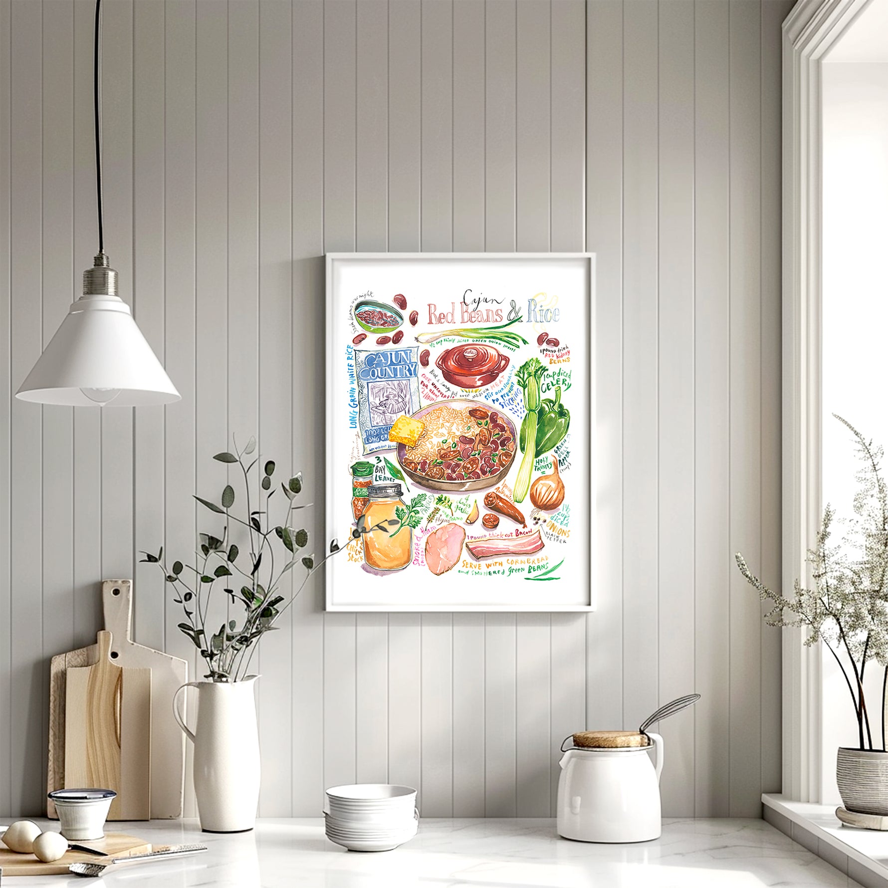 cajun red beans and rice recipe fine art print in watercolor featuring the ingredients and the finished dish, along with handwritten instructions in English. The framed poster hanging in a kitchen space.