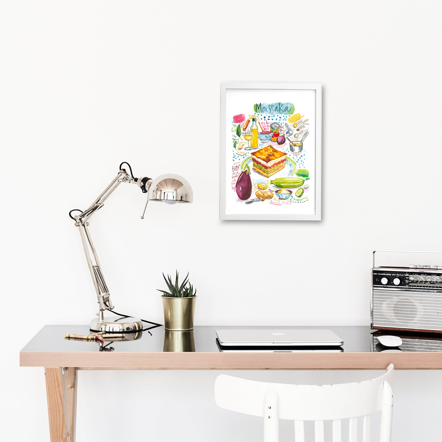moussaka recipe fine art print in watercolor featuring the ingredients and the finished dish, along with handwritten instructions in English. The framed poster hanging in a working space.