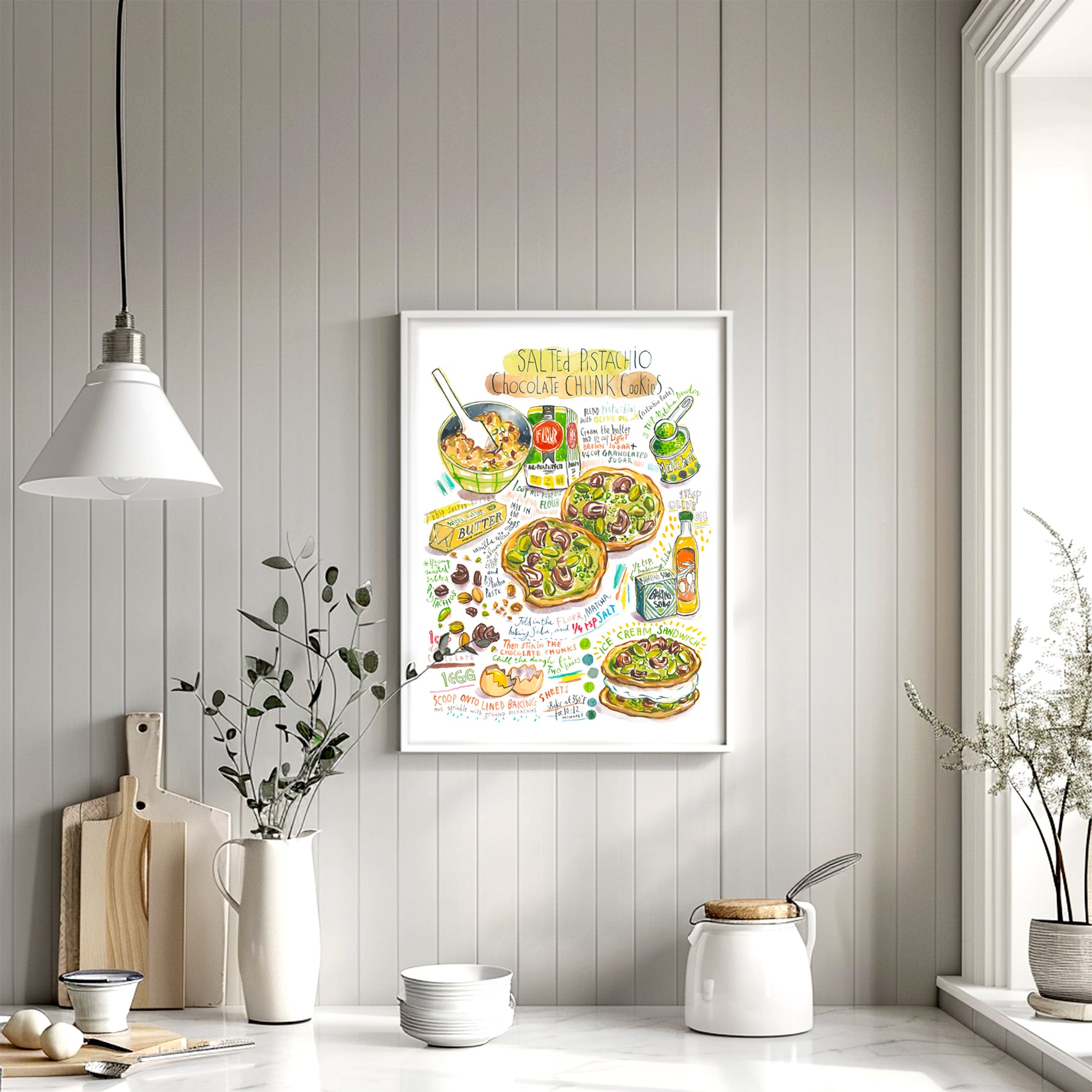 Salted pistachio chocolate chunk cookies recipe fine art print in watercolor featuring the ingredients and two cookies, along with handwritten instructions in English. The framed poster hanging in a dining space.